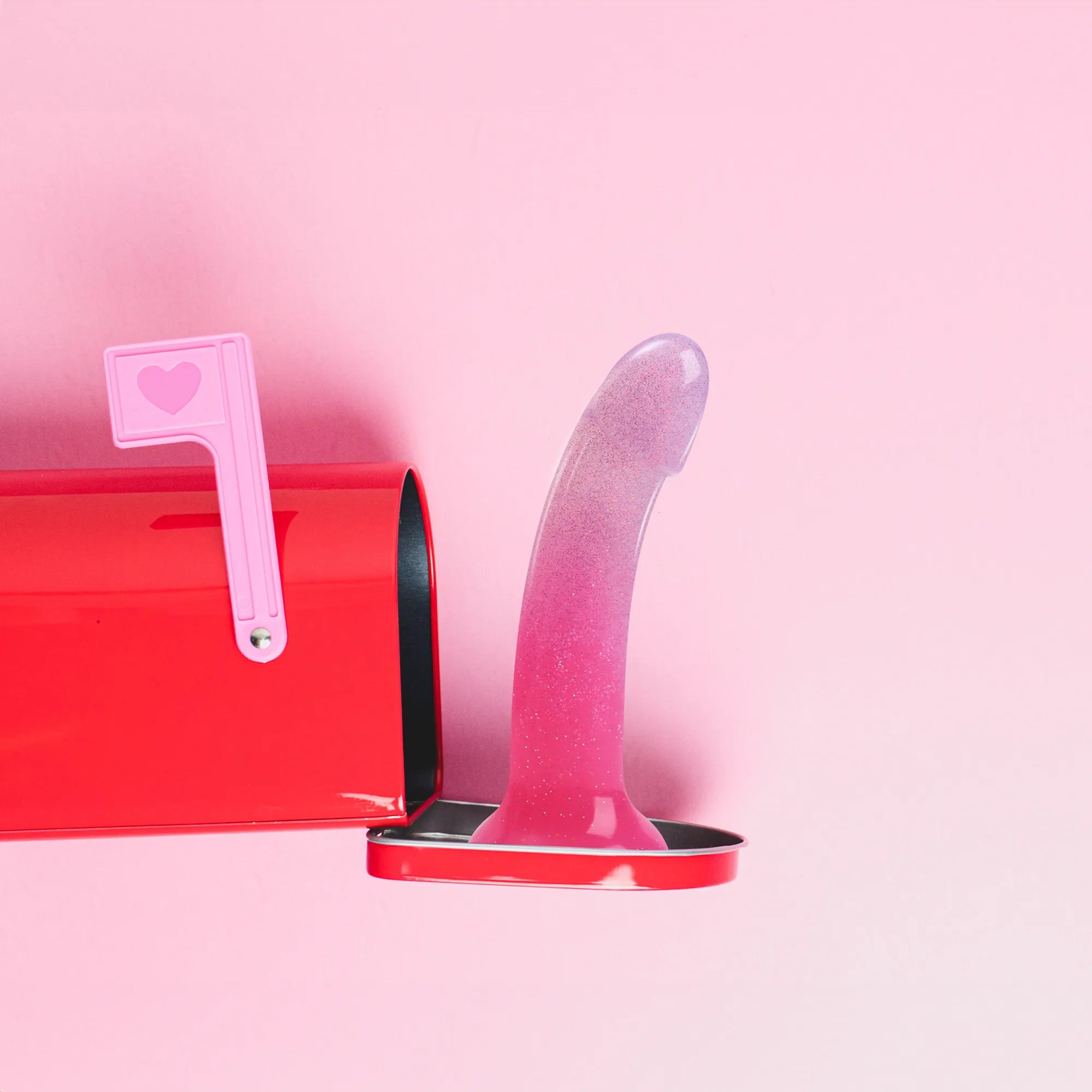 Gode design en silicone DILDOLLS rose translucide avec effet pailleté, mis en scène avec une boîte aux lettres rouge