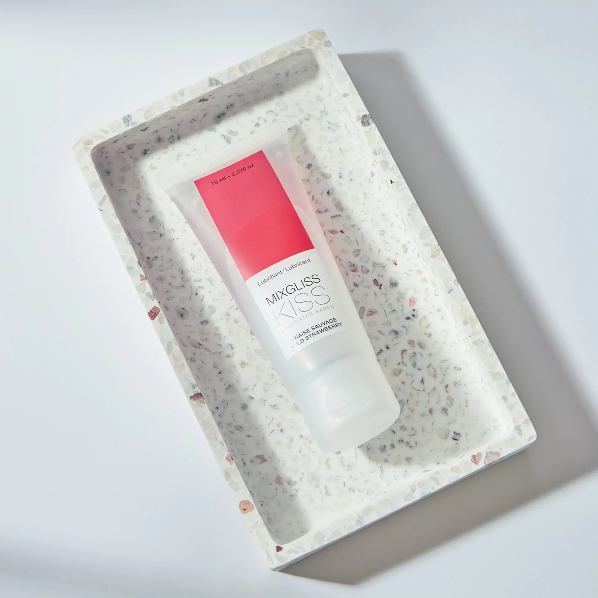 Tube de lubrifiant Mixgliss Kiss fraise posé dans un cadre terrazzo – produit intime au design épuré