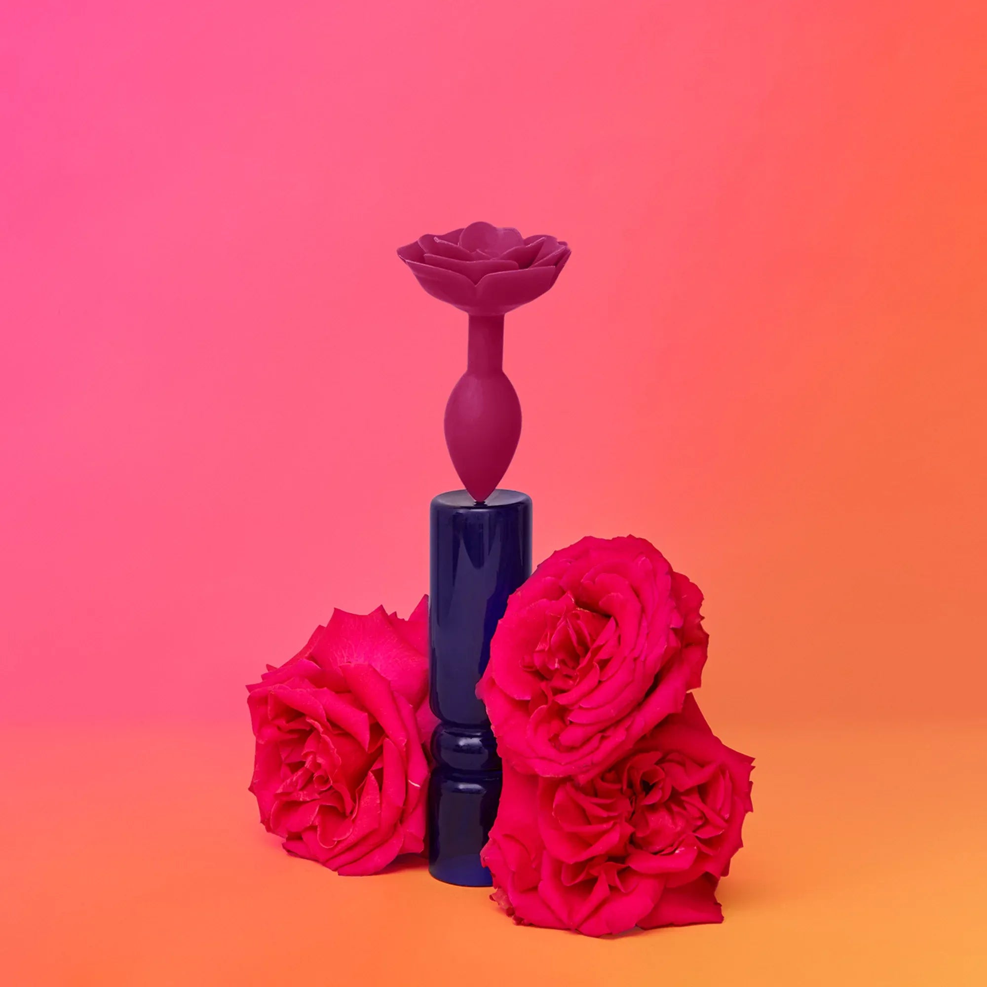 Plug anal en silicone rose foncé Open Roses posé sur un socle bleu avec des roses rouges sur fond rose orangé